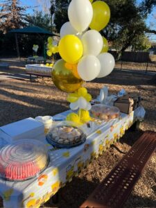 Outdoor birthday party catering setup with festive yellow and white balloons, assorted pastries, and beverages arranged on a decorated picnic table. 