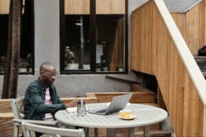A man sits alone at an outdoor café table with a coffee and laptop, surrounded by a quiet, impersonal setting. Despite the sunny atmosphere, the scene conveys a sense of isolation, capturing Adrian’s early experiences in Portola Valley, where chain cafés lacked the warmth, creativity, and community he once cherished.